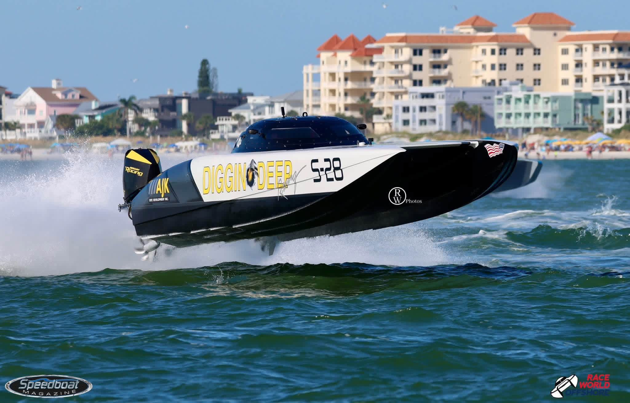 Diggin Deep S-28 airborne over a wave, with beachside buildings behind the race boat.